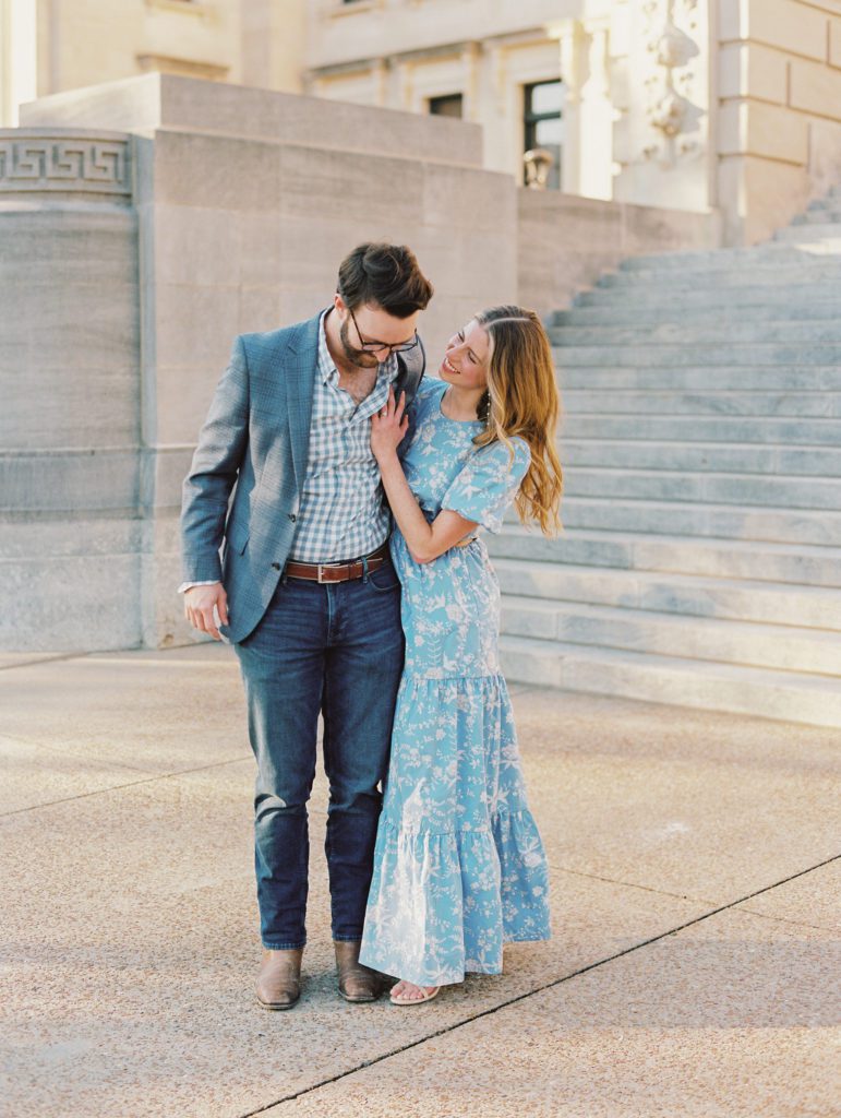 Engaged couple taking photos on the steps of City Hall, Jackson Mississippi wearing a french blue cut out maxi dress and a grey suit with a gingham shirt.