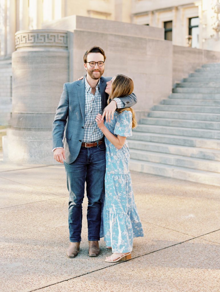 Sweet engaged couple taking photos on the steps of City Hall, Jackson Mississippi wearing a french blue cut out maxi dress and a grey suit with a gingham shirt.