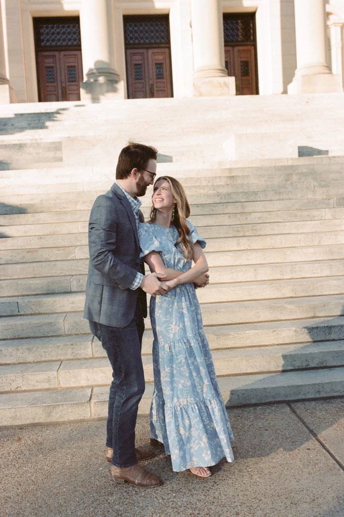 Engaged couple taking photos on the steps of City Hall, Jackson Mississippi wearing a french blue cut out maxi dress and a grey suit with a gingham shirt.